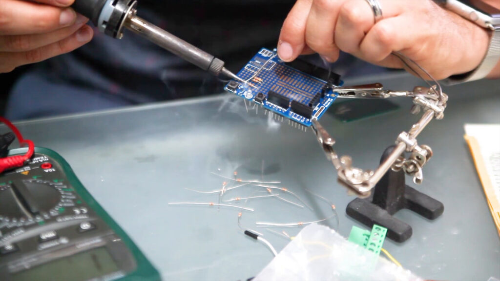 Close-up of hands soldering a blue circuit board in a helping-hands clamp, a reference image from Ivan Guaderrama's talk